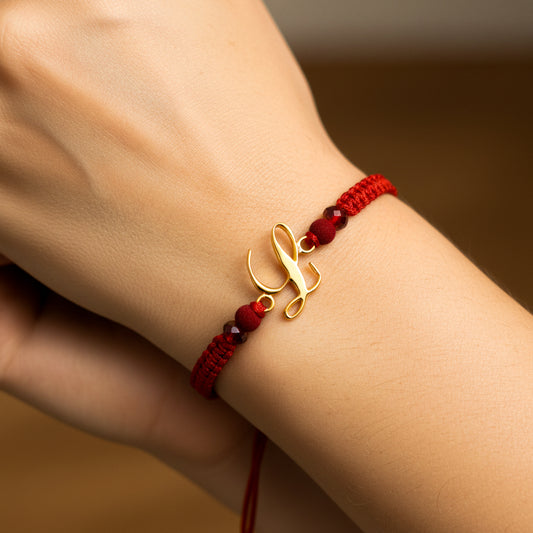 Mano de mujer usando una pulsera tejida en hilo de color rojo con bisutería del mismo color con herraje de la letra L en oro amarillo de 18 quilates de Joyería Caracas. 