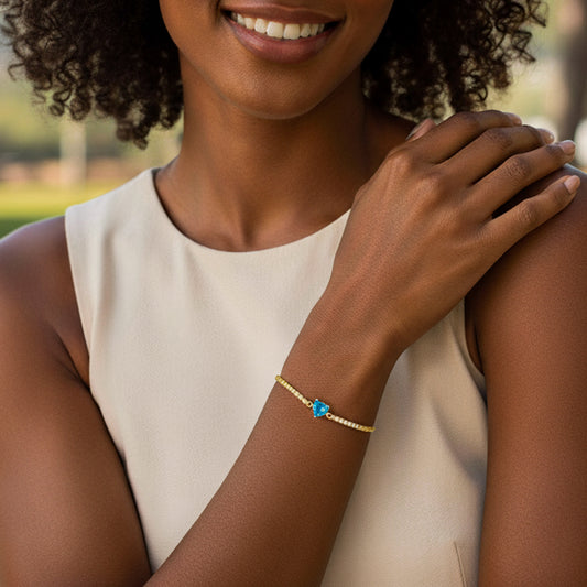 Mano de mujer usando una pulsera en oro amarillo de 18 quilates con cristales de color blanco en los laterales y un cristal de color azul claro en forma de corazón en el centro de Joyería Caracas. 