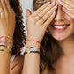 Mano de mujer con pulsera tejida en hilo, con accesorios de color verde y dije de San Benito en oro amarillo de 18 quilates, hecho en Joyería Caracas. 