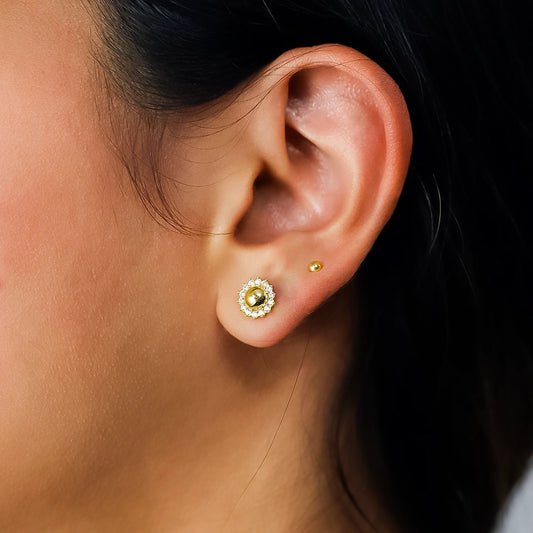 Oreja de mujer usando un topito en forma de girasol en oro amarillo de 18 quilates con cristales de color blanco alrededor en Joyería Caracas. 