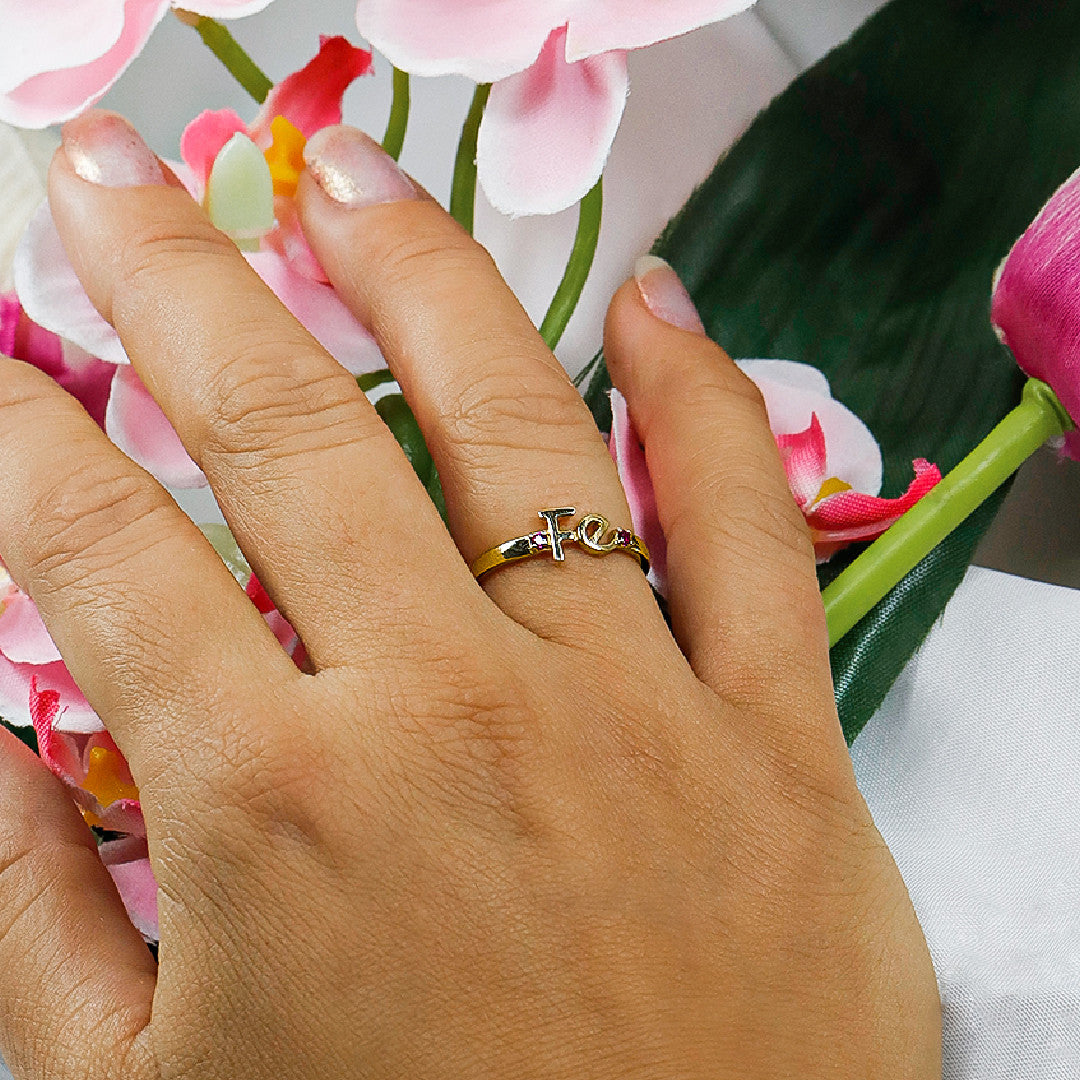 Mano de mujer con anillo en forma de la palabra fe en oro amarillo de 18 quilates con cristales de color rosa, hecho en Joyería Caracas. 