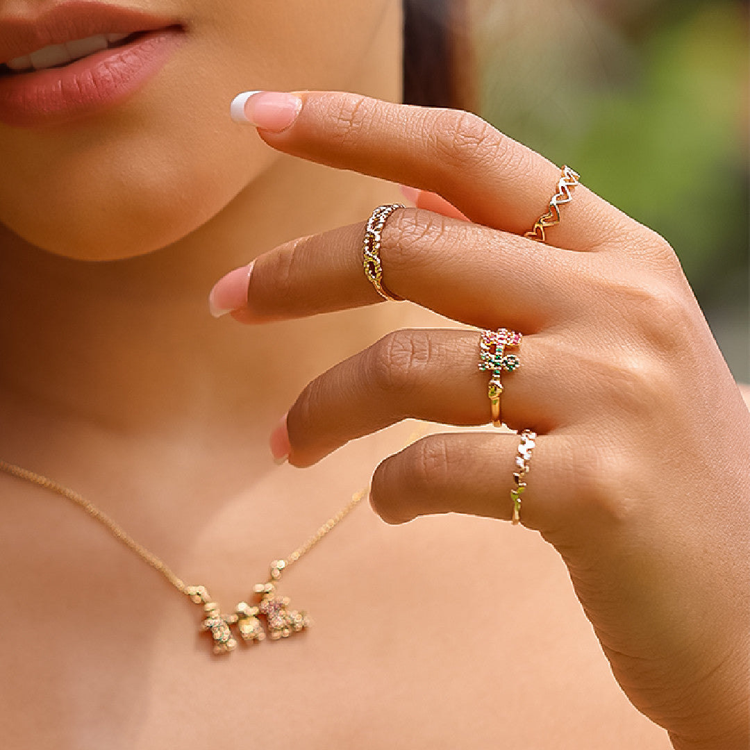 Mano de mujer usando anillo con forma de niño y niña en oro amarillo de 18 quilates con cristales de color verde y rosa, hecho en Joyería Caracas. 