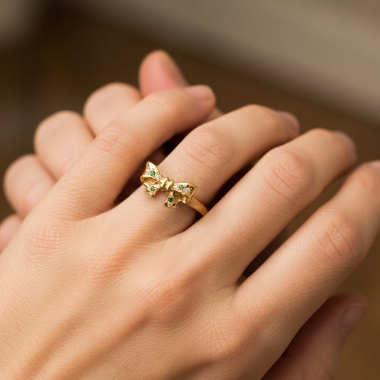 Mano de mujer usando un anillo con la forma de un corbatín en el centro en oro amarillo de 18 quilates con cristales de color verde y blanco de Joyería Caracas. 