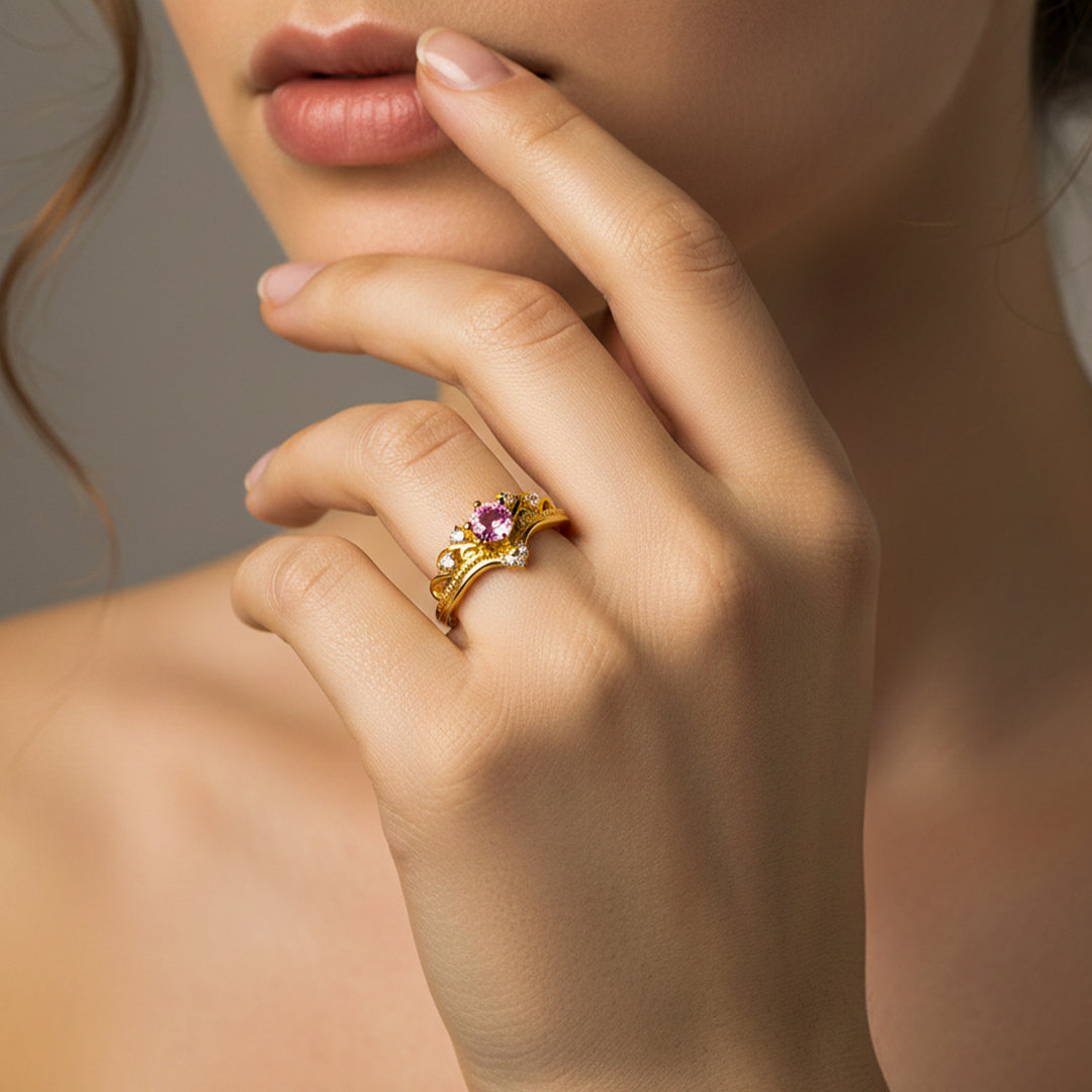 Mano de mujer usando un anillo en forma de corona inglesa en oro amarillo de 18 quilates con cristales de color blanco y 1 rosa en el centro de Joyería Caracas. 