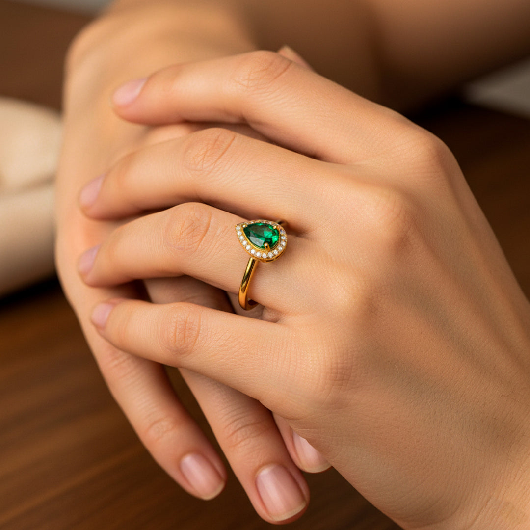 Mano de mujer usando un anillo en oro amarillo de 18 quilates con la forma de una gota en el centro con cristal en forma de gota de color verde y cristales blancos alrededor de Joyería Caracas. 