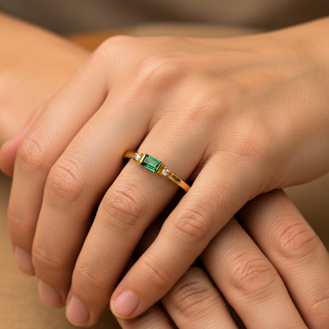 Mano de mujer usando un anillo en oro amarillo de 18 quilates con Esmeralda en el centro y 2 cristales de color blanco de Joyería Caracas. 