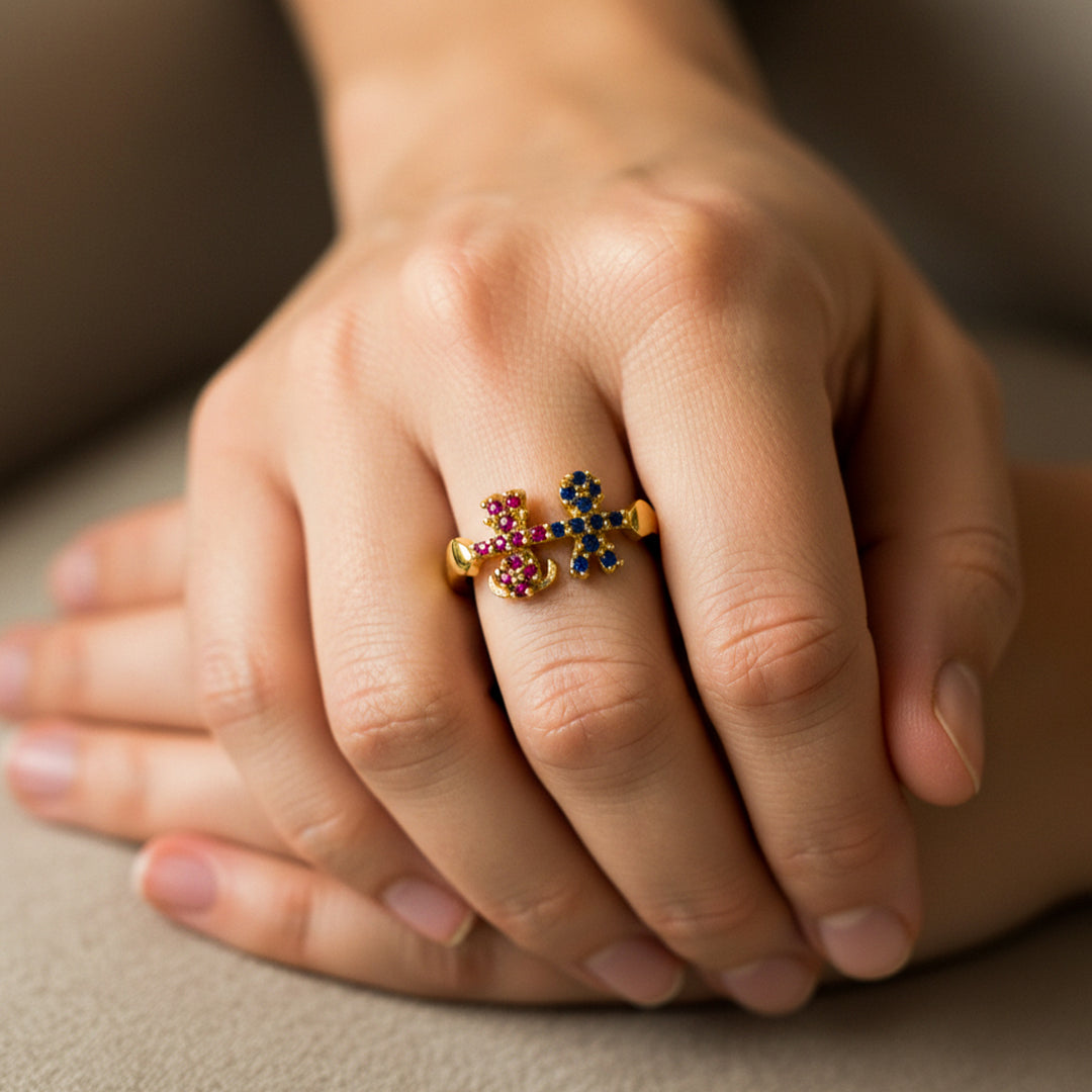 Mano de mujer usando un anillo en oro amarillo de 18 quilates con la forma de un niño y niña tomados de la mano con cristales de color azul y rosa de Joyería Caracas. 