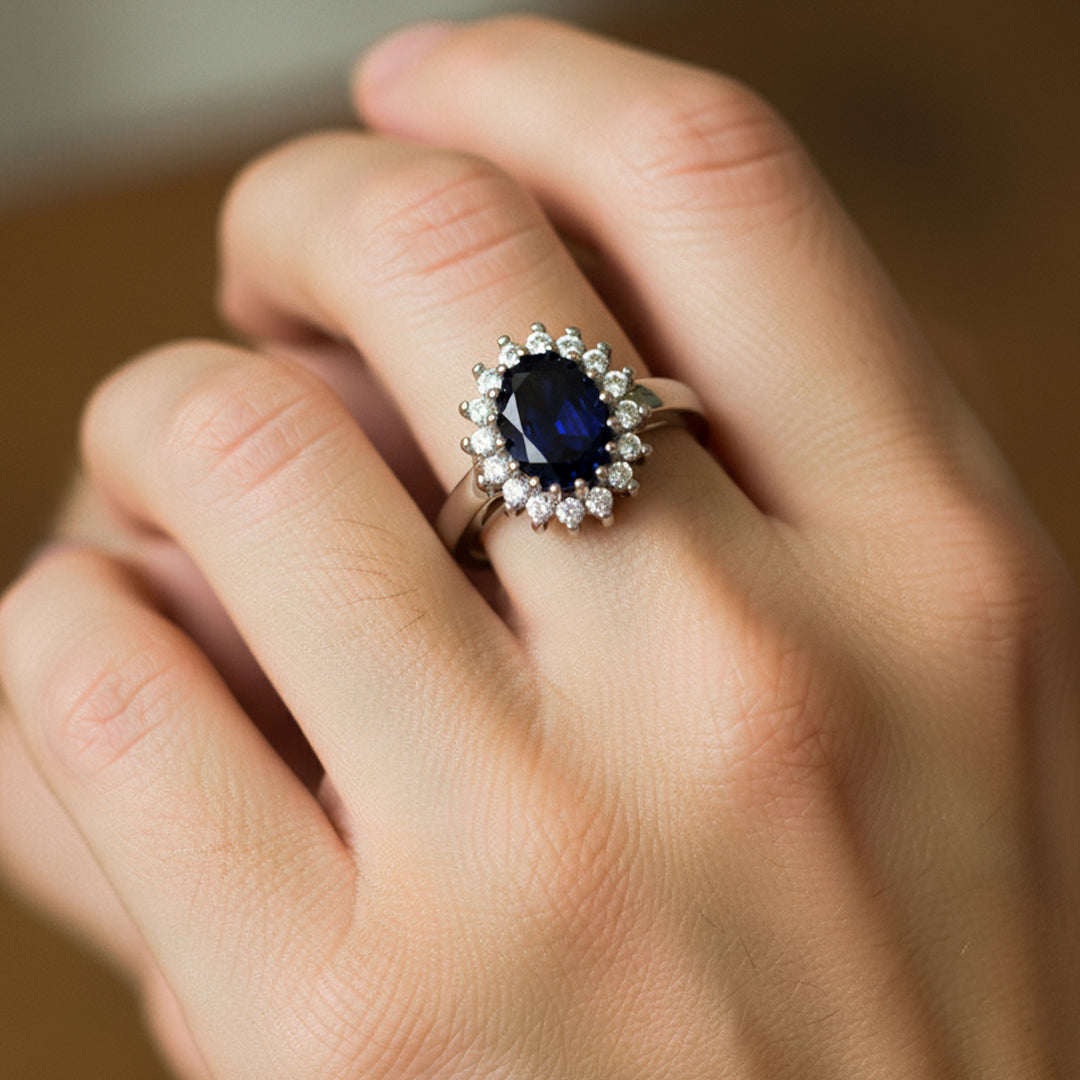 Mano de mujer usando un anillo diseño Lady Di en oro blanco de 18 quilates con cristal azul ovalado en el centro y alrededor blancos de Joyería Caracas. 