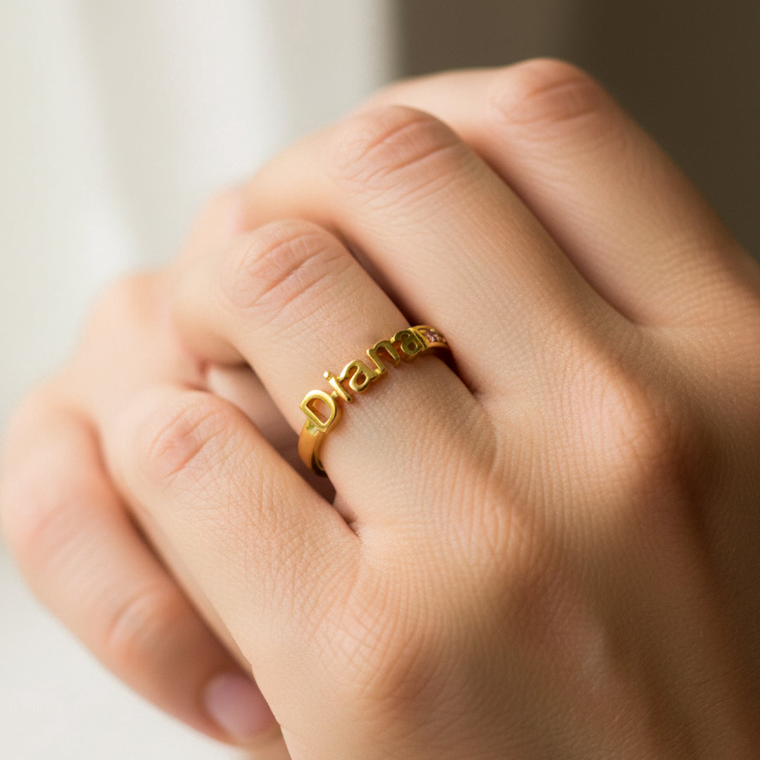Mano de mujer usando un anillo de nombre personalizado en oro amarillo de 18 quilates con cristales de color lila de Joyería Caracas. 