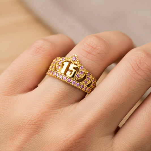Mano de mujer usando un anillo en forma de corona en oro amarillo de 18 quilates con el numero 15 en el centro y cristales de color rosa alrededor de Joyería Caracas. 