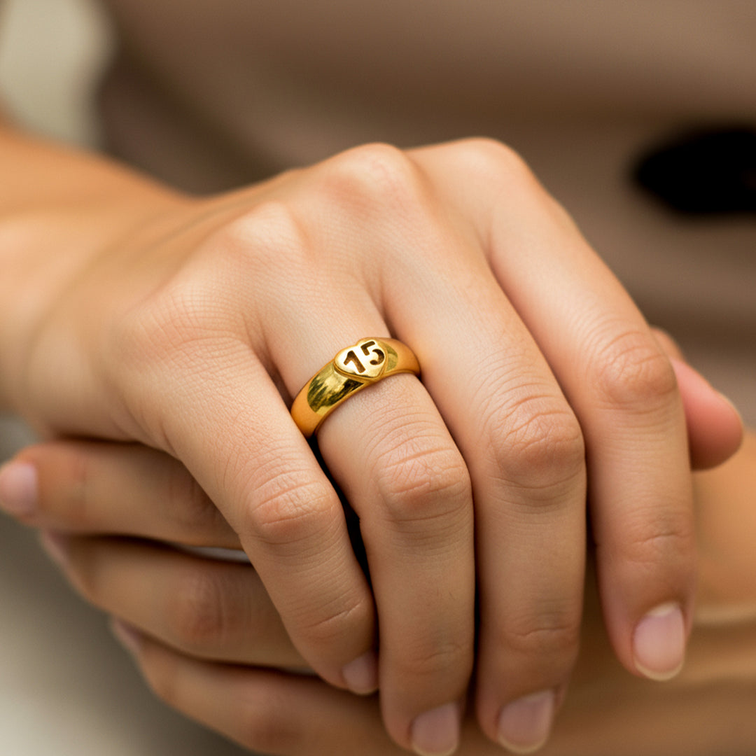 Mano de mujer usando un anillo plano con el número 15 dentro de un corazón en el centro en oro amarillo de 18 quilates de Joyería Caracas. 