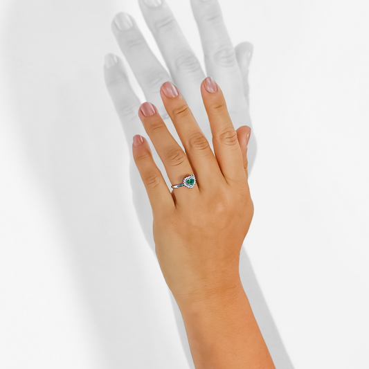 Mano de mujer usando anillo con forma de corazón en el centro en oro blanco de 18 quilates con piedra esmeralda de color verde en forma de corazón y 20 diamantes de color blanco, hecho en Joyería Caracas. 