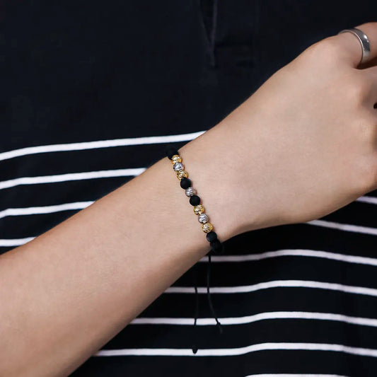 Mano de hombre usando una pulsera tejida en hilo de color negro con accesorios del mismo color y esferas facetadas en oro amarillo y blanco de 18 quilates de Joyería Caracas. 