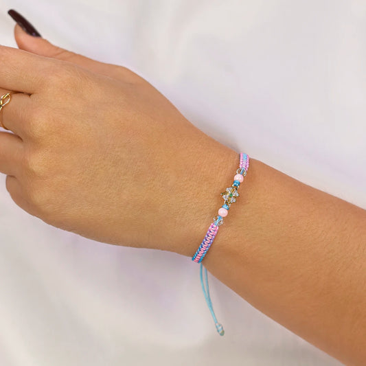 Mano de mujer con pulsera tejida en hilo de distintos colores, accesorios de bisutería de color blanco, rosa y dije en forma de trébol en oro amarillo de 18 quilates con cristales de color blanco y verde, hecho en Joyería Caracas. 