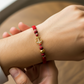 Mano de modelo usando una pulsera tejida de hilo color rojo, herraje en forma de ancla en oro amarillo de 18 quilates con cristales de color blanco, esfera facetada y bisutería de calidad de Joyería Caracas. 