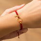 Mano de modelo usando una pulsera tejida de hilo color rojo con dije circular, 2 esferas lisas y dije de la letra A en oro amarillo de 18 quilates de Joyería Caracas. 