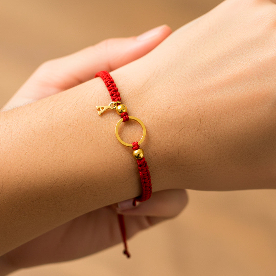 Mano de modelo usando una pulsera tejida de hilo color rojo con dije circular, 2 esferas lisas y dije de la letra A en oro amarillo de 18 quilates de Joyería Caracas. 