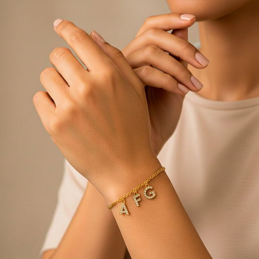 Mano de mujer usando una pulsera en tejido bismarck con dijes de iniciales de la letra A, F Y G en oro amarillo de 18 quilates con cristales de color blanco de Joyería Caracas. 