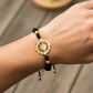 Mano de modelo usando una pulsera tejida en hilo color negro con herraje en forma de brújula y esferas facetadas en oro amarillo de 18 quilates de Joyería Caracas. 