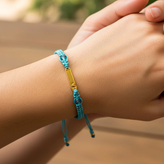 Mano de modelo usando una pulsera tejida en hilo color celeste, con placa lisa con corazón en oro amarillo de 18 quilates y bisutería de calidad de Joyería Caracas. 