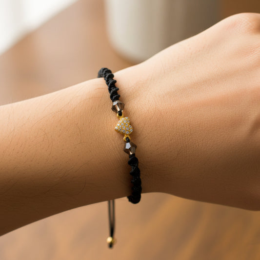Mano de modelo usando una pulsera tejida en hilo color negro con dije en forma de corazón en oro amarillo de 18 quilates con cristales de color blanco y bisutería de calidad de Joyería Caracas. 