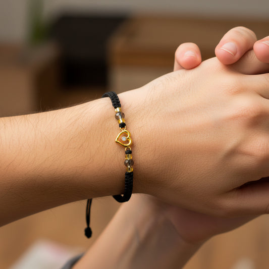 Mano de modelo usando una pulsera tejida en hilo color negro con dije en forma de corazón en oro amarillo de 18 quilates con cristal de color blanco y bisutería de calidad de Joyería Caracas. 