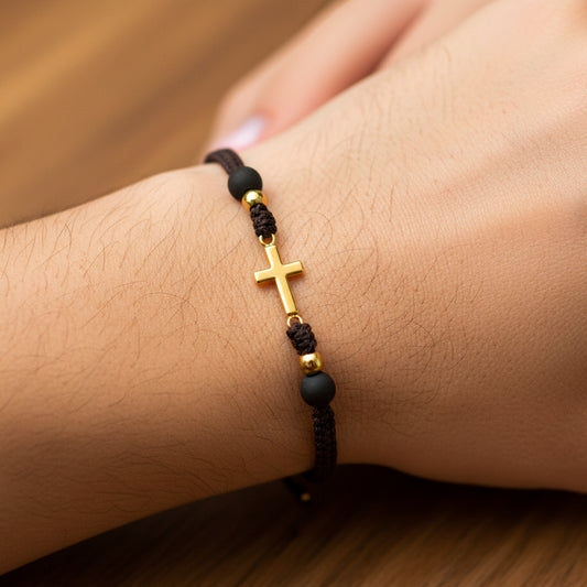 Mano de modelo usando una pulsera tejida en hilo color negro con dije de cruz y esferas lisas en oro amarillo de 18 quilates con bisutería de calidad de Joyería Caracas. 
