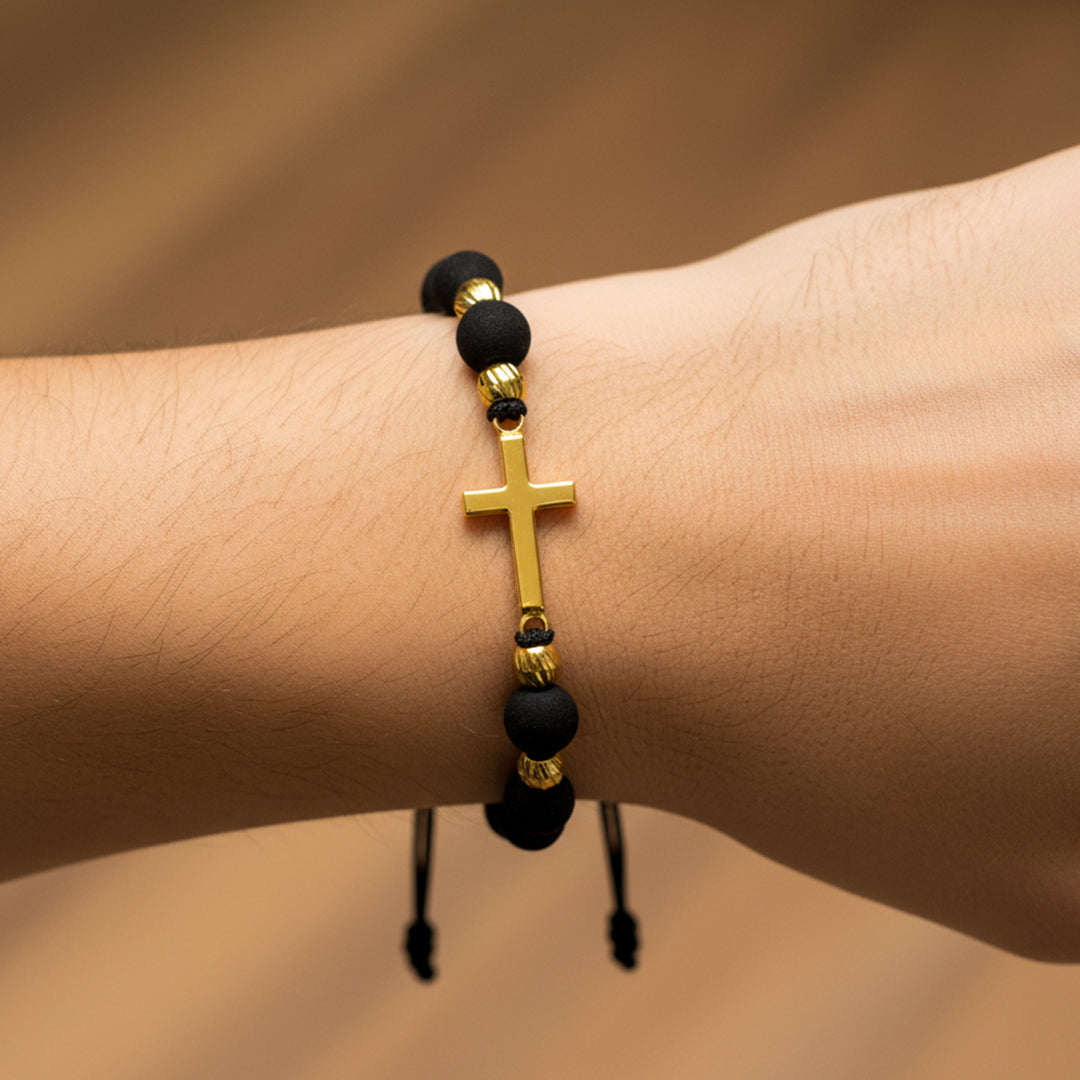 Mano de modelo usando una pulsera tejida en hilo de color negro con dije de cruz con esferas facetadas en oro amarillo de 18 quilates con bisutería de calidad de Joyería Caracas. 