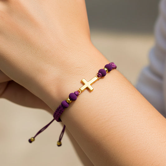Mano de modelo usando una pulsera tejida en hilo color morado con dije de cruz y esferas lisas en oro amarillo de 18 quilates de Joyería Caracas. 