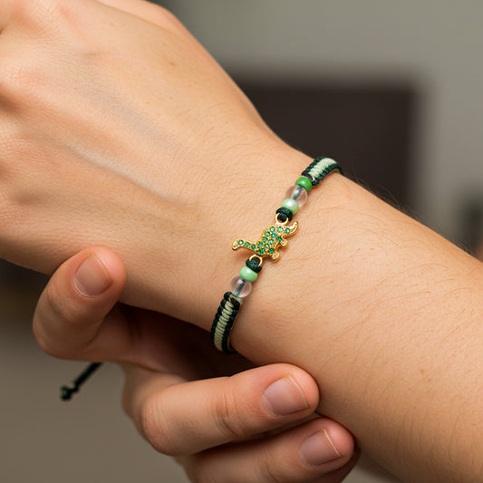 Mano de modelo usando una pulsera tejida de hilo color verde, herraje en forma de dinosaurio en oro amarillo de 18 quilates con cristales de color verde y bisutería de calidad de Joyería Caracas. 