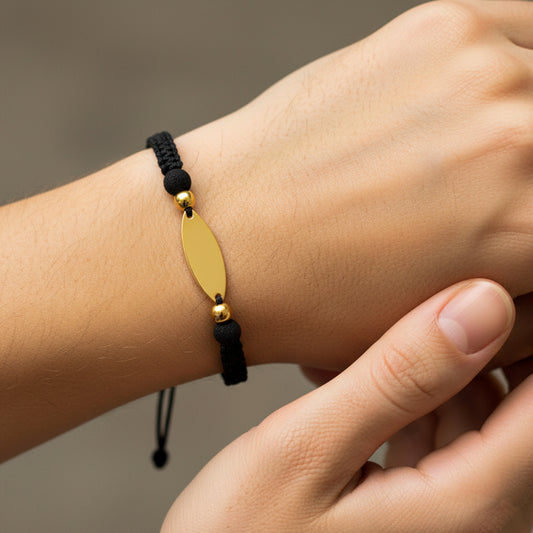 Mano de modelo usando una pulsera tejida con hilo de color negro y placa ovalada con 2 esferas lisas en oro amarillo de 18 quilates y bisutería de calidad de Joyería Caracas. 