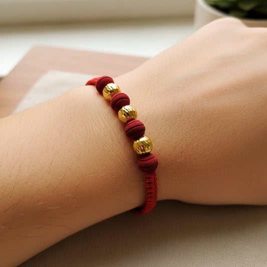 Mano de modelo usando una pulsera tejida en hilo color rojo, con 3 esferas facetadas en oro amarillo de 18 quilates y bisutería de calidad de Joyería Caracas. 