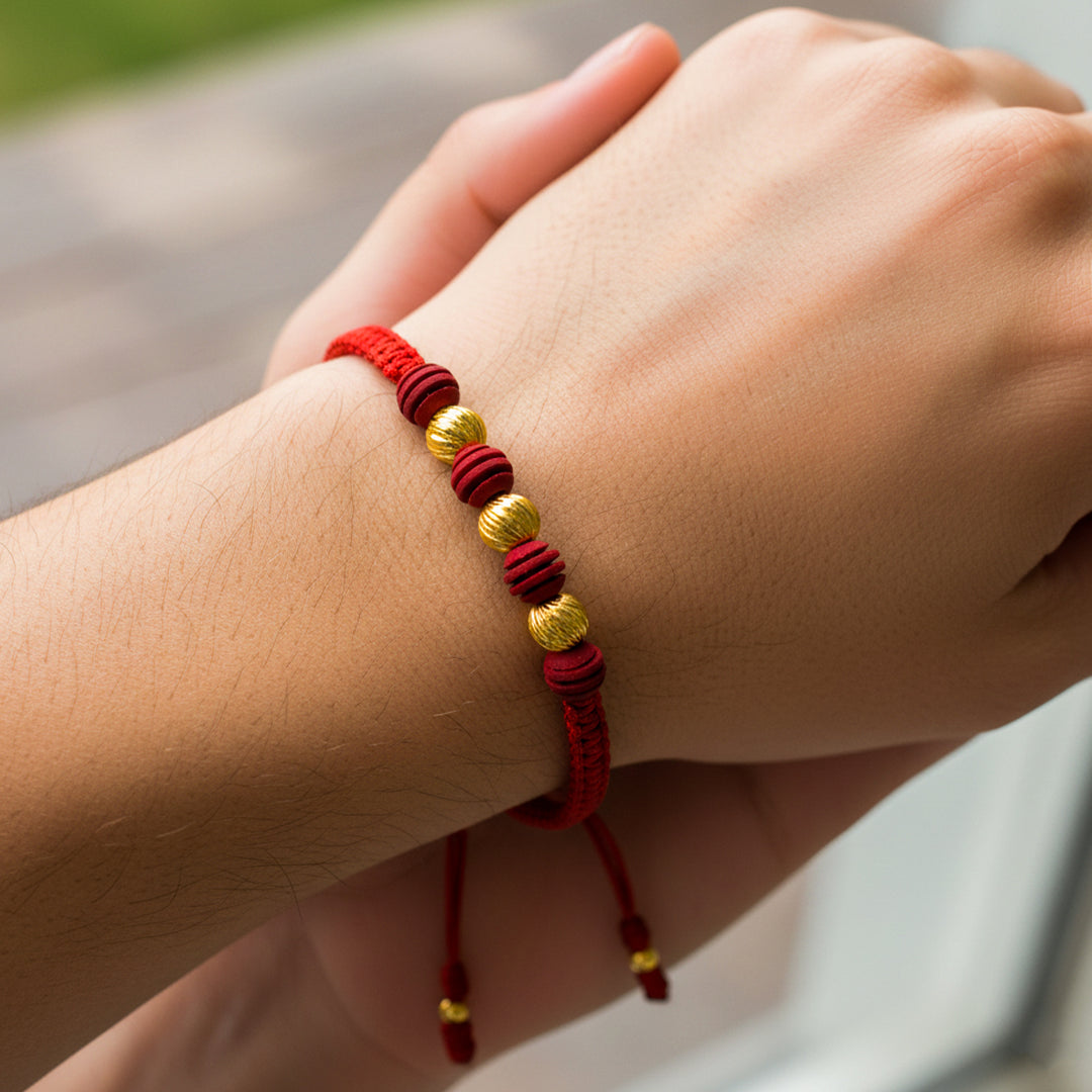 Mano de mujer usando una pulsera tejida en hilo de color rojo, bisutería del mismo color y 3 esferas facetadas en oro amarillo de 18 quilates de Joyería Caracas. 