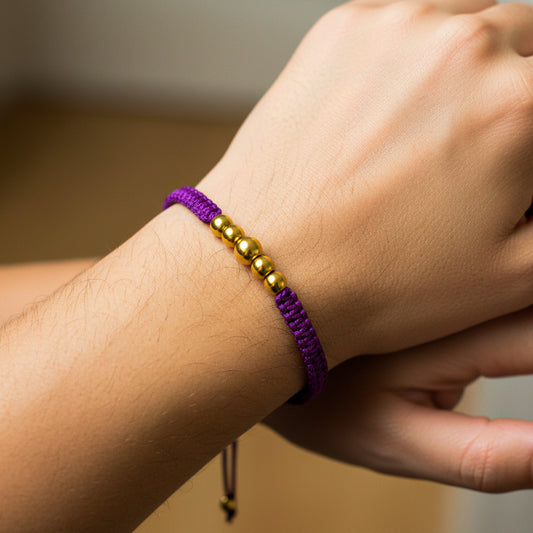 Mano de modelo usando una pulsera tejida en hilo de color morado con esferas lisas en oro amarillo de 18 quilates de Joyería Caracas. 