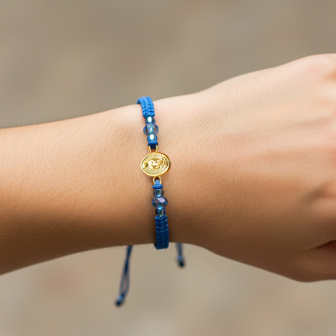 Mano de modelo usando una pulsera tejida en hilo color celeste con dije de la Virgen de Guadalupe en oro amarillo de 18 quilates de Joyería Caracas. 