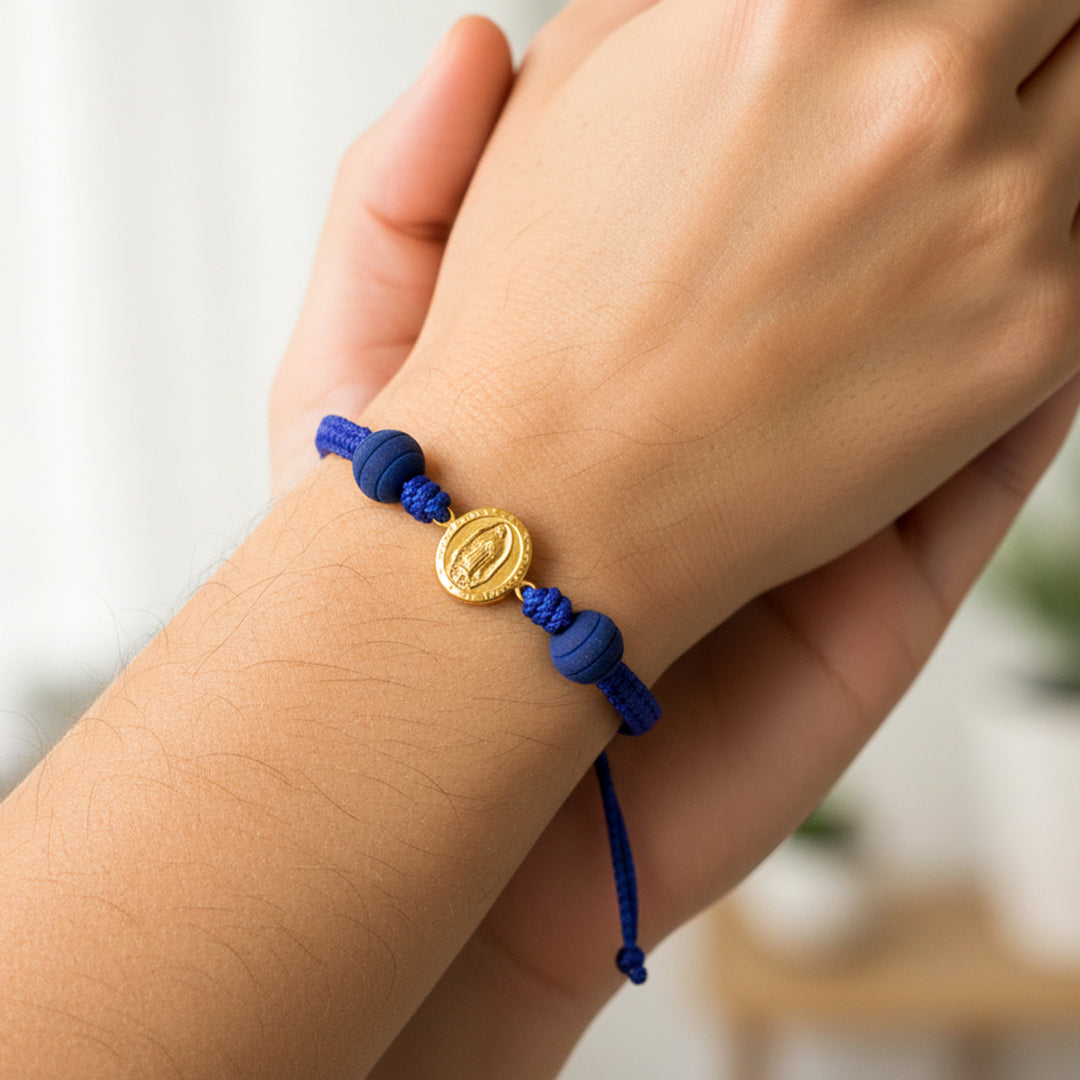 Mano de modelo usando una pulsera tejida en hilo color azul oscuro con dije de la Virgen de Guadalupe en oro amarillo de 18 quilates y bisutería de calidad de Joyería Caracas.