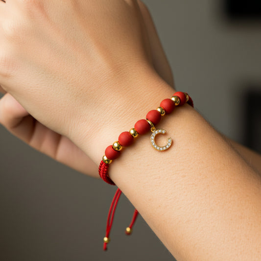 Mano de modelo usando una pulsera tejida en hilo color rojo con dije de la letra C y esferas lisas en oro amarillo de 18 quilates con cristales de color blanco y bisutería de calidad de Joyería Caracas. 