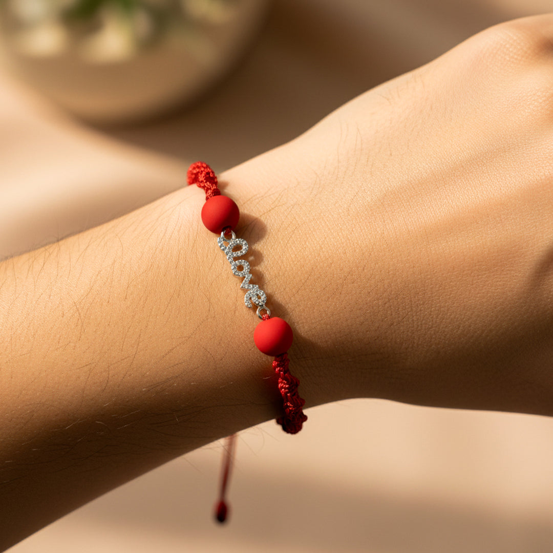 Mano de modelo usando una pulsera tejida en hilo de color rojo con un dije de la palabra love en oro blanco de 18 quilates con cristales de color blanco y bisutería de calidad de Joyería Caracas. 