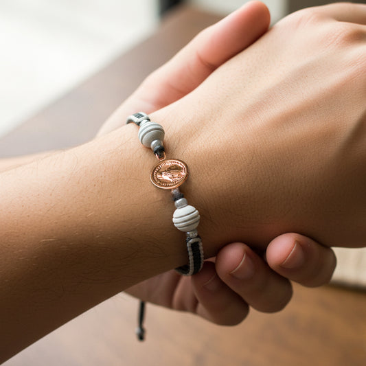 Mano de modelo usando una pulsera tejida en hilo color gris con dije de la Virgen milagrosa en oro rosa de 18 quilates  de Joyería Caracas.