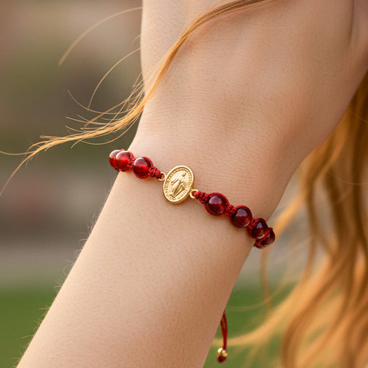 Mano de modelo usando una pulsera tejida en hilo color rojo con accesorios color rojo y herraje de la Virgen Milagrosa en oro amarillo de 18 quilates de Joyería Caracas. 