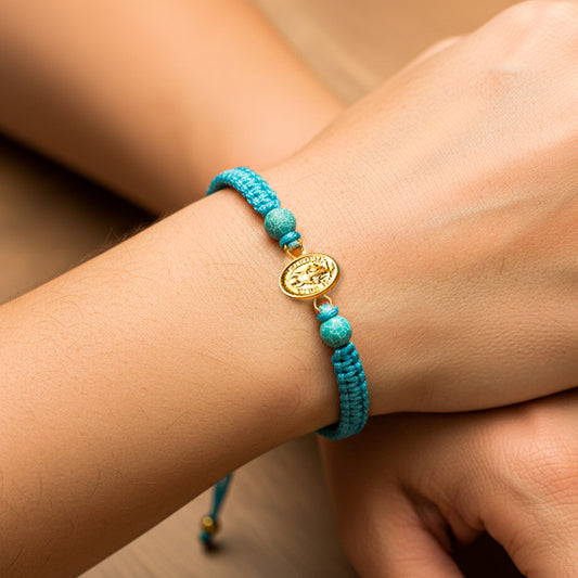 Mano de modelo usando una pulsera tejida en hilo color celeste con dije de la milagrosa en oro amarillo de 18 quilates de Joyería Caracas. 