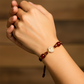 Mano de modelo usando una pulsera tejida en hilo color vinotinto con dije de la milagrosa en oro amarillo de 18 quilates con cristales de color blanco y bisutería de calidad de Joyería Caracas.