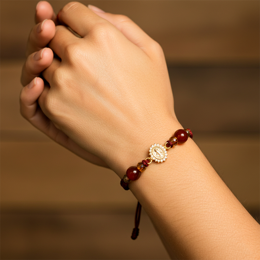 Mano de modelo usando una pulsera tejida en hilo color vinotinto con dije de la milagrosa en oro amarillo de 18 quilates con cristales de color blanco y bisutería de calidad de Joyería Caracas.