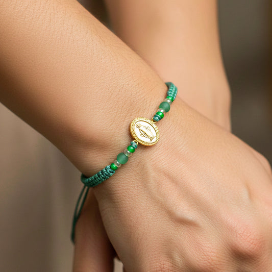 Mano de modelo usando una pulsera tejida en hilo color verde aguamarina con accesorios y herraje de la Virgen Milagrosa en oro amarillo de 18 quilates de Joyería Caracas. 
