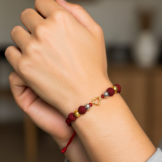 Mano de modelo usando una pulsera tejida en hilo de color rojo con dije en forma de corazón y esferas facetadas en oro amarillo y blanco de 18 quilates con bisutería de calidad de Joyería Caracas. 