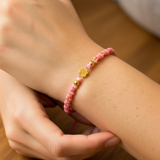 Mano de modelo usando una pulsera tejida en hilo color rosa con herraje en forma de osito con 2 esferas lisas en oro amarillo de 18 quilates y bisutería de calidad de Joyería Caracas. 