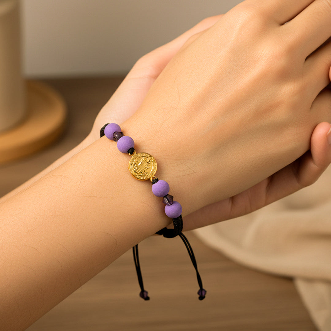Mano de modelo usando una pulsera tejida con hilo de color negro, dije de San Benito en oro amarillo de 18 quilates y bisutería de calidad de Joyería Caracas. 
