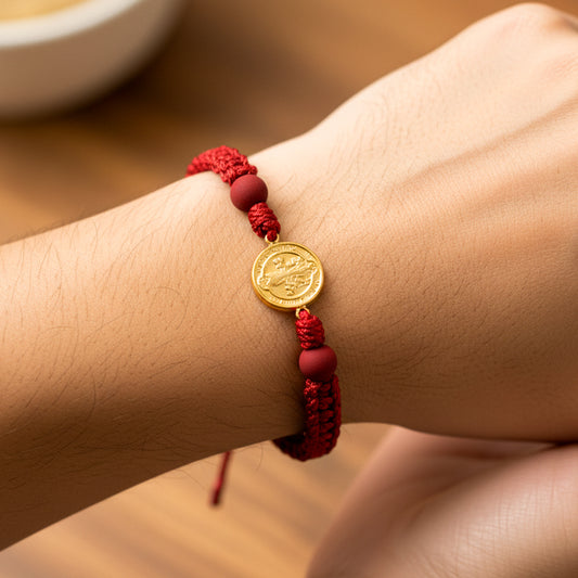 Mano de modelo usando una pulsera tejida en hilo de color rojo con dije de San Benito en oro amarillo de 18 quilates de Joyería Caracas. 