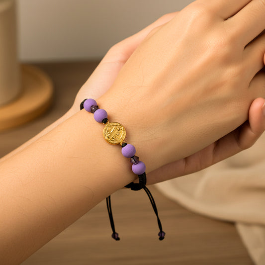 Mano de modelo usando una pulsera tejida con hilo de color negro, dije de San Benito en oro amarillo de 18 quilates y bisutería de calidad de Joyería Caracas. 