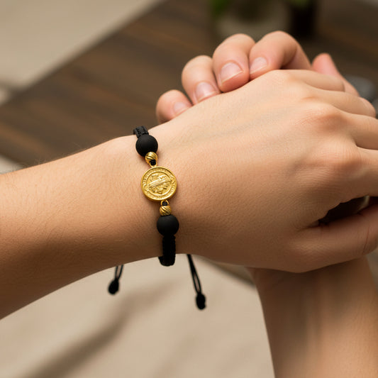 Mano de modelo usando una pulsera tejida en hilo color negro con 2 esferas facetadas, dije de San Benito en oro amarillo de 18 quilates con bisutería de calidad de Joyería Caracas. 